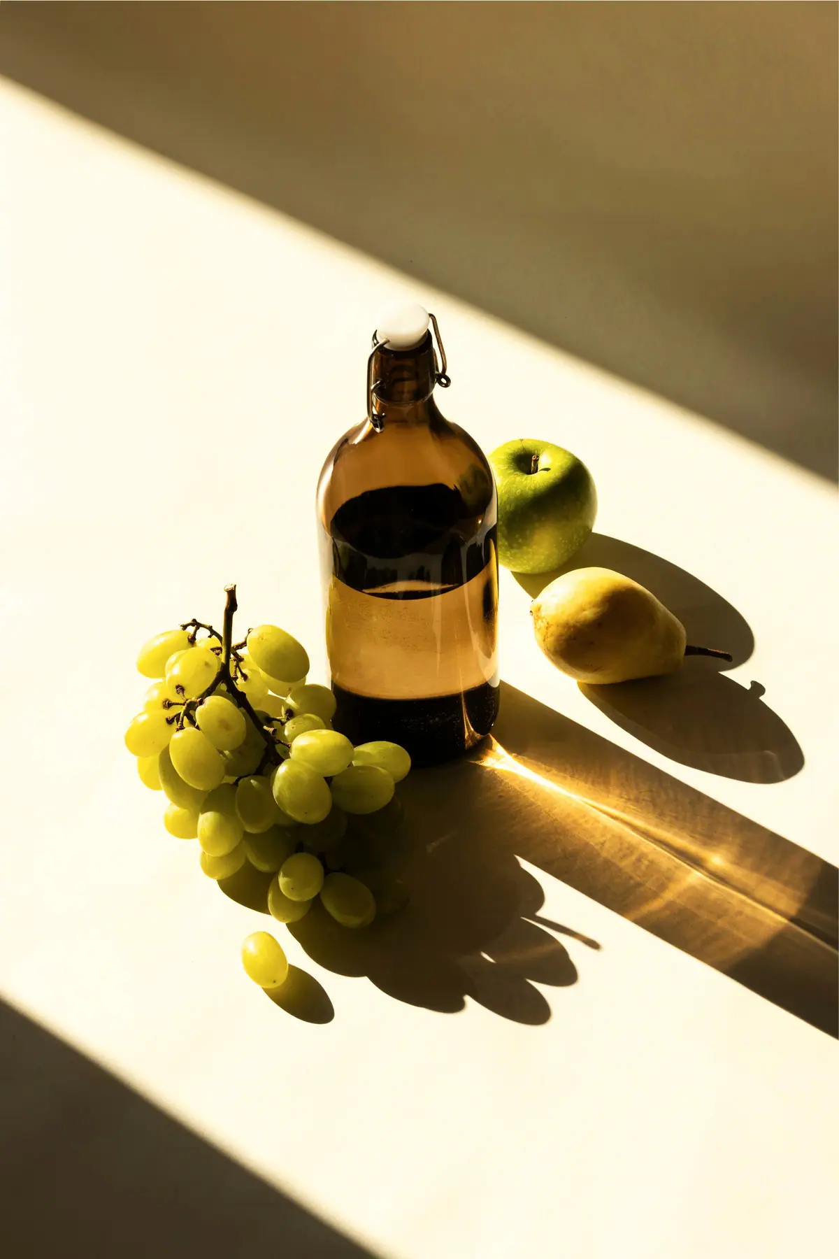 An amber glass bottle with green grapes and pears on a pale ledge, in hard afternoon light