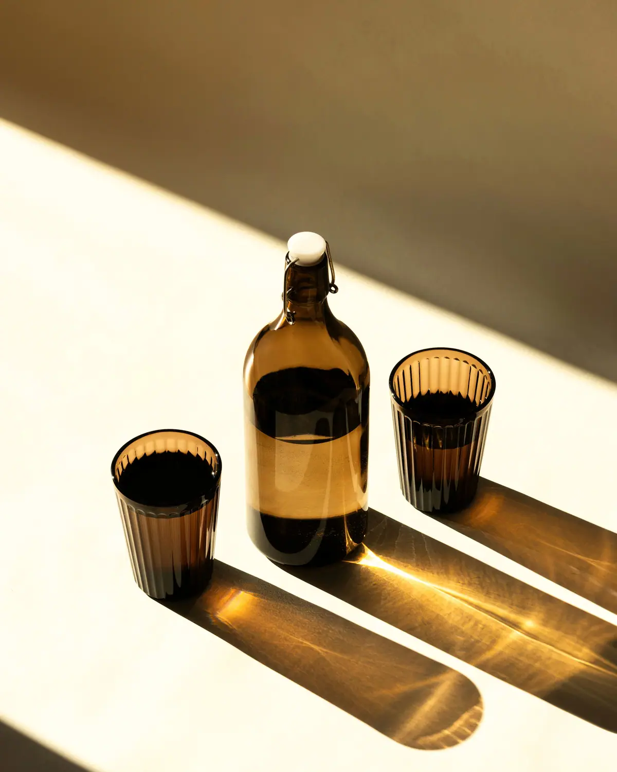 An amber glass bottle and two brown glasses casting long hard shadows across a white surface in low sun