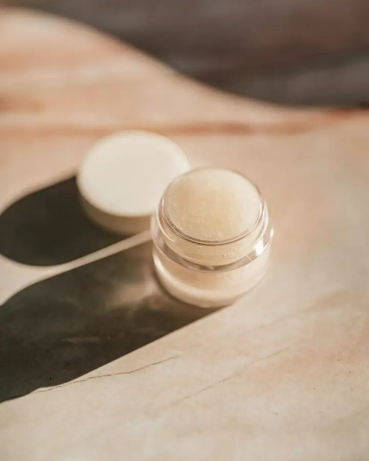 Small frosted glass pot of balm with its white lid beside it, in hard sunlight on a warm beige surface