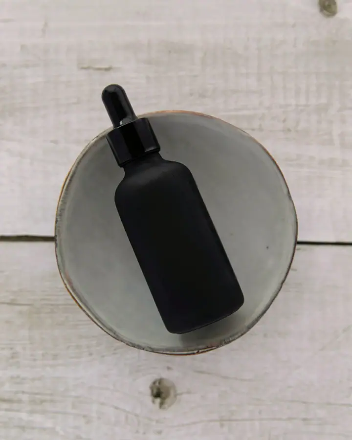 Matte black glass dropper bottle lying on a round stone plate, seen from above